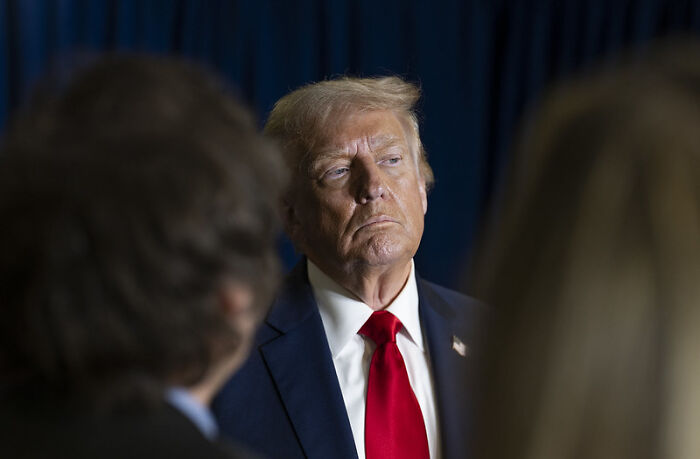 Man in a suit with a red tie standing among a crowd, appearing deep in thought during a public event.
