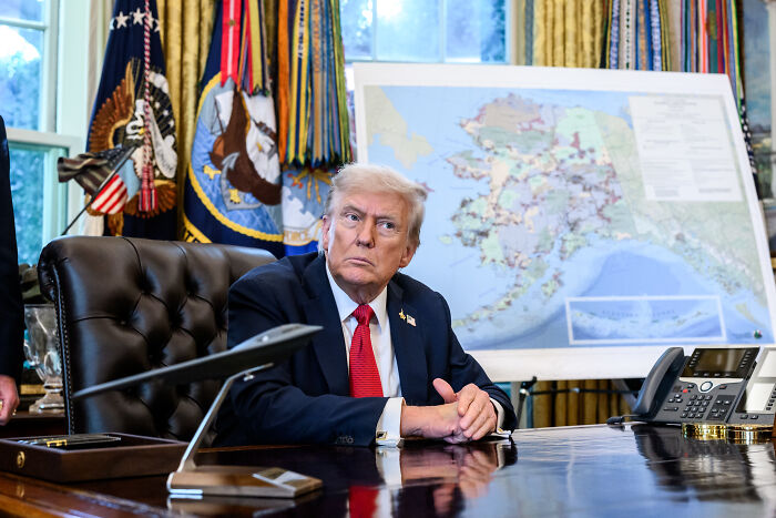 Donald Trump sitting at the desk in the Oval Office with a map of Alaska in the background during a formal meeting. Donald Trump sitting at the desk in the Oval Office with a map of Alaska in the background during a formal meeting.