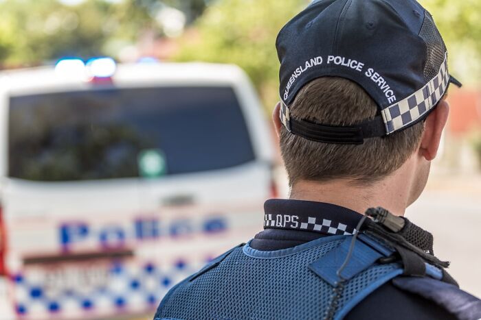 Police officer from Queensland Police Service facing away, standing near a police van at an outdoor scene. Police officer from Queensland Police Service facing away, standing near a police van at an outdoor scene.