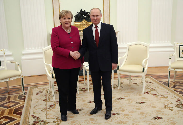 Angela Merkel shaking hands with Vladimir Putin in a formal room, related to Poland and Baltic States fueling Putin’s war.