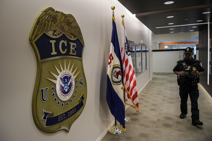 ICE emblem on wall with US and agency flags, a uniformed officer walking down a hallway inside a government facility. ICE emblem on wall with US and agency flags, a uniformed officer walking down a hallway inside a government facility.