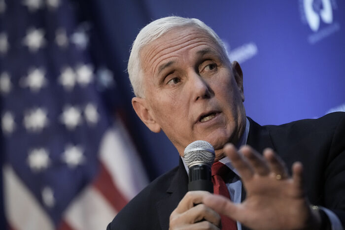 Mike Pence speaking at an event, holding a microphone, with a blurred American flag background, related to Trump called Pence wimp. Mike Pence speaking at an event, holding a microphone, with a blurred American flag background, related to Trump called Pence wimp.