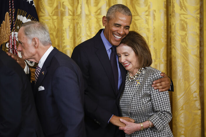 Barack Obama warmly hugging Nancy Pelosi in a formal setting with golden curtains in the background. Barack Obama warmly hugging Nancy Pelosi in a formal setting with golden curtains in the background.