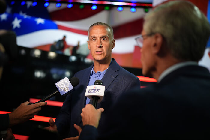 A Trump adviser speaking to reporters with microphones, discussing ICE agents at Bad Bunny’s Super Bowl performance.