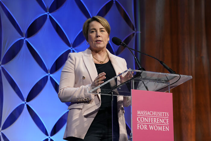Aide to Gov Maura Healey speaking at Massachusetts Conference for Women event, wearing a light blazer at a clear podium. Aide to Gov Maura Healey speaking at Massachusetts Conference for Women event, wearing a light blazer at a clear podium.