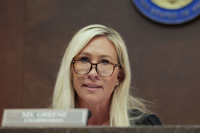 Marjorie Taylor Greene speaking in a formal setting, wearing glasses and seated before a microphone. Marjorie Taylor Greene speaking in a formal setting, wearing glasses and seated before a microphone.