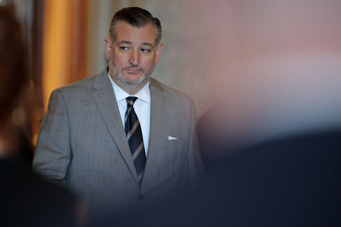 Ted Cruz in a gray suit and striped tie, looking to the side during a formal event with blurred people in foreground. Ted Cruz in a gray suit and striped tie, looking to the side during a formal event with blurred people in foreground.