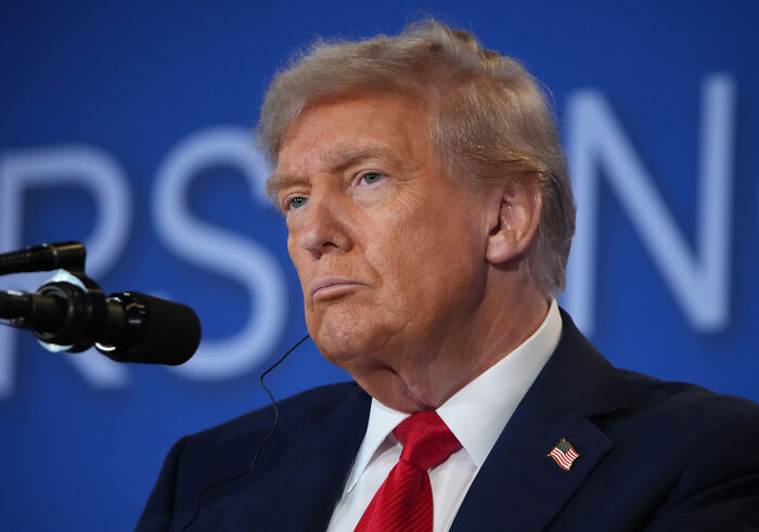Donald Trump speaking at a microphone, dressed in suit and red tie, with blue background featuring blurred text. Donald Trump speaking at a microphone, dressed in suit and red tie, with blue background featuring blurred text.