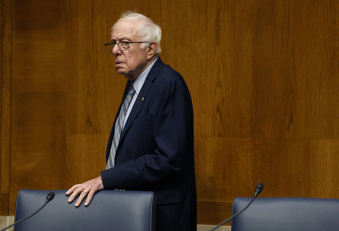 Bernie Sanders standing in a navy suit and striped tie in a wood-paneled room during a formal event. Bernie Sanders standing in a navy suit and striped tie in a wood-paneled room during a formal event.