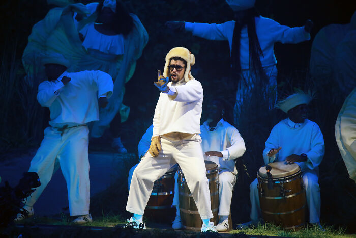 Performer in white outfit and hat dancing on stage with drummers, related to MAGA and Turning Point USA poster controversy.