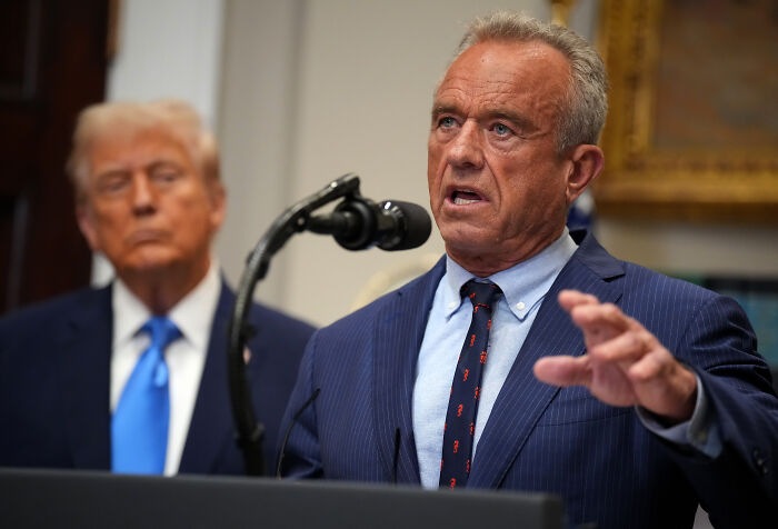 RFK Jr. speaking at a podium with a microphone in a suit, addressing infectious disease official removal.