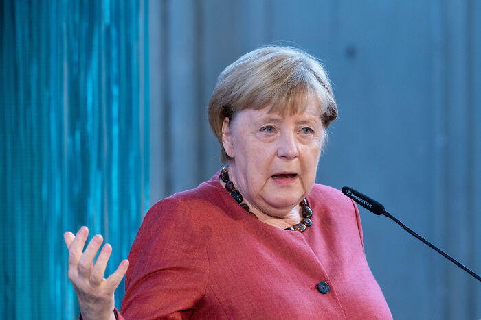 Angela Merkel speaking at a podium, discussing Poland and Baltic states fueling Putin’s war in Ukraine.