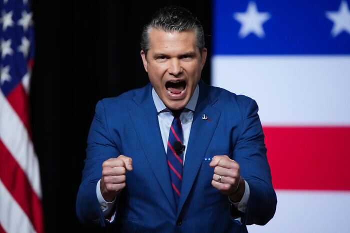 Man in a blue suit speaking passionately with clenched fists in front of a blurred American flag backdrop. Man in a blue suit speaking passionately with clenched fists in front of a blurred American flag backdrop.