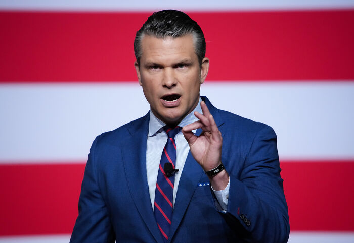 Man in blue suit and striped tie speaking emphatically in front of an American flag backdrop about fat troops remarks.