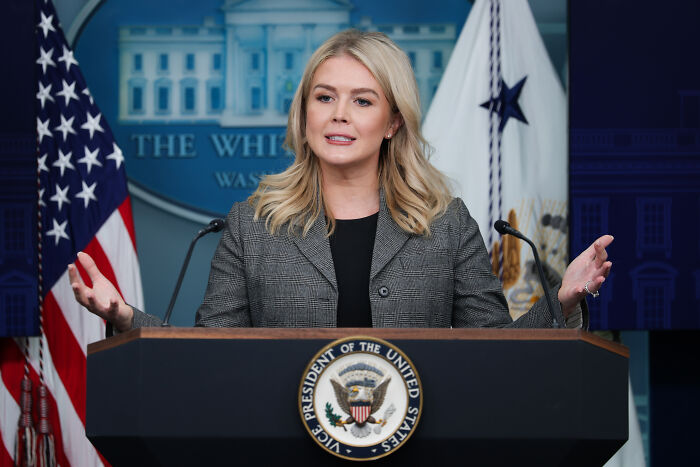 Karoline Leavitt speaking at a podium with US flags behind her, defending Trump’s AI medbed video.