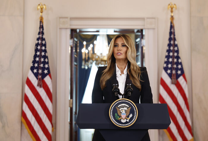 Melania Trump standing at a podium with presidential seal, flanked by American flags in an official setting.