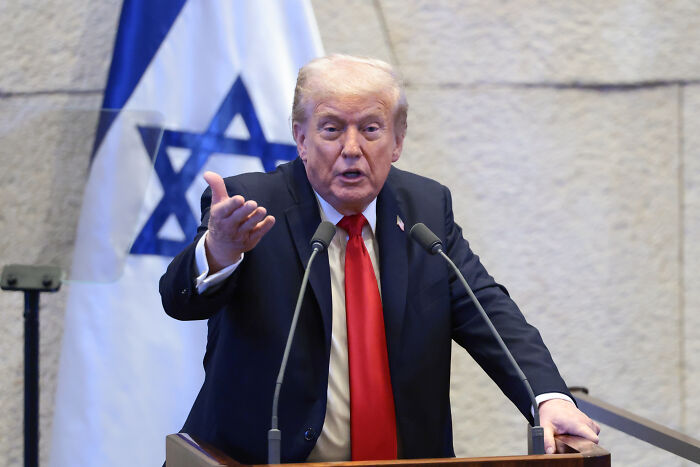 Donald Trump speaking at a podium with Israeli flag behind him during a Knesset event.
