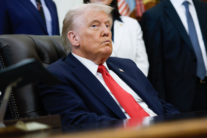 Trump sitting in a suit and red tie with a serious expression, linked to a sinister clip about his presidency continuing indefinitely.