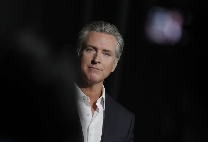 Gavin Newsom wearing a dark suit and white shirt, speaking at an event with a serious expression on a dark background Gavin Newsom wearing a dark suit and white shirt, speaking at an event with a serious expression on a dark background