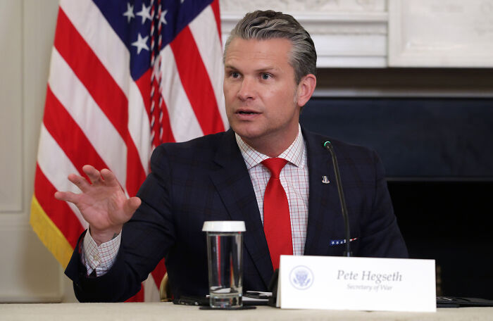 Pete Hegseth speaking at a briefing, wearing a dark suit and red tie, with an American flag in the background. Pete Hegseth speaking at a briefing, wearing a dark suit and red tie, with an American flag in the background.