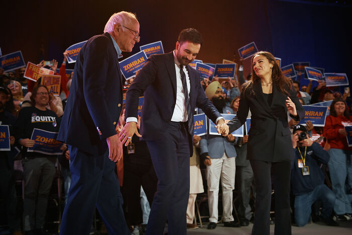 AOC at a campaign event holding hands with two men, with supporters holding signs in the background. AOC at a campaign event holding hands with two men, with supporters holding signs in the background.