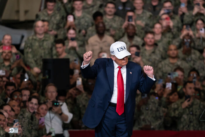 Donald Trump wearing a USA hat and red tie, gesturing during a speech aboard an aircraft carrier with military personnel watching. Donald Trump wearing a USA hat and red tie, gesturing during a speech aboard an aircraft carrier with military personnel watching.