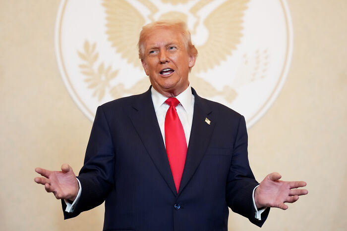 Donald Trump in a dark suit with a red tie speaking and gesturing during an Asia summit event with a presidential emblem backdrop. Donald Trump in a dark suit with a red tie speaking and gesturing during an Asia summit event with a presidential emblem backdrop.