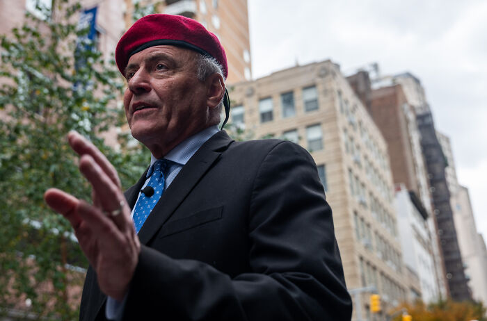 Man in a red beret and suit speaking outdoors in a city setting, related to NYC mayoral frontrunner Zohran Mamdani controversy.