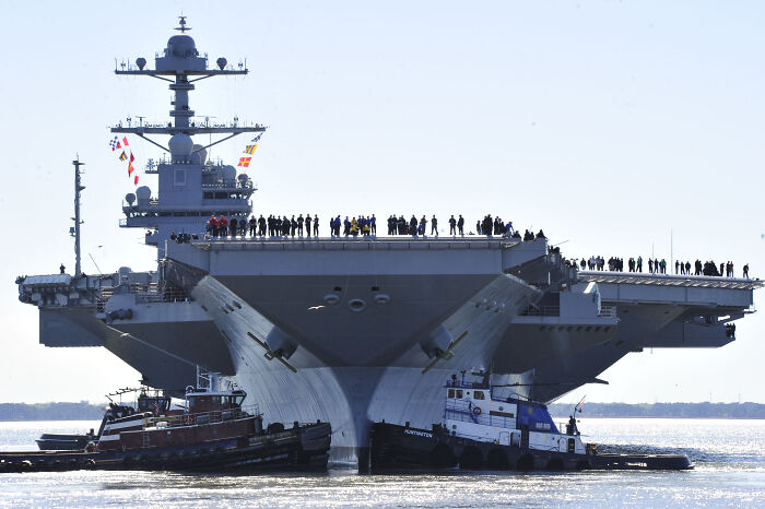 U.S. Navy’s largest aircraft carrier sailing with crew on deck, escorted by tugboats in open water near the Caribbean. U.S. Navy’s largest aircraft carrier sailing with crew on deck, escorted by tugboats in open water near the Caribbean.