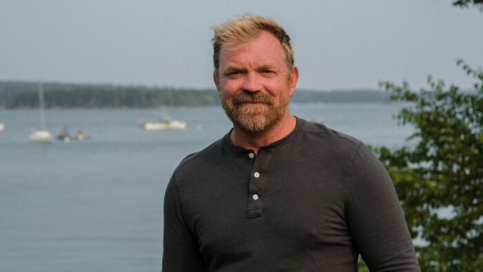 Maine Senate candidate outdoors by water, wearing a dark long-sleeve shirt, covering tattoo recognized as hate symbol.