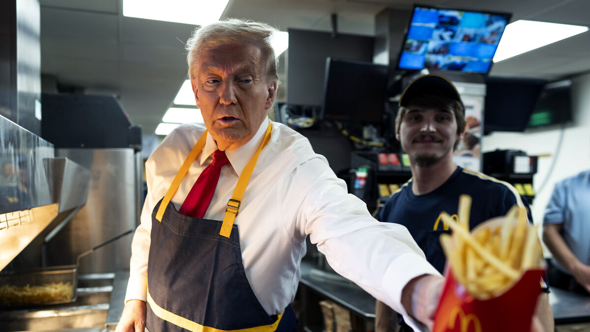 Donald Trump serving French fries wearing an apron in a fast food kitchen with a worker in the background.