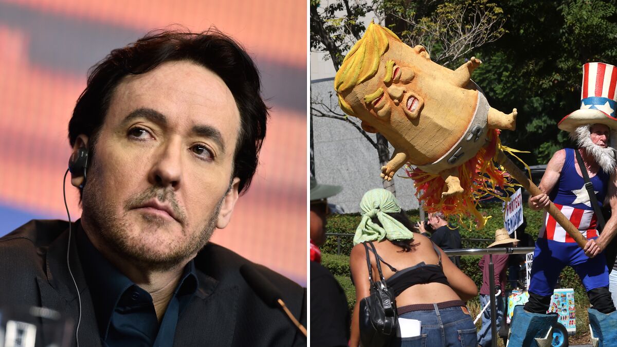 John Cusack wearing earpiece speaking at event next to a protester holding a Trump piu00f1ata with an Uncle Sam costume.