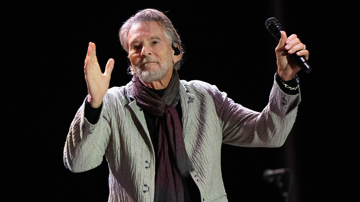 Singer of Danger Zone performing on stage, holding a microphone and wearing a gray jacket with a scarf.
