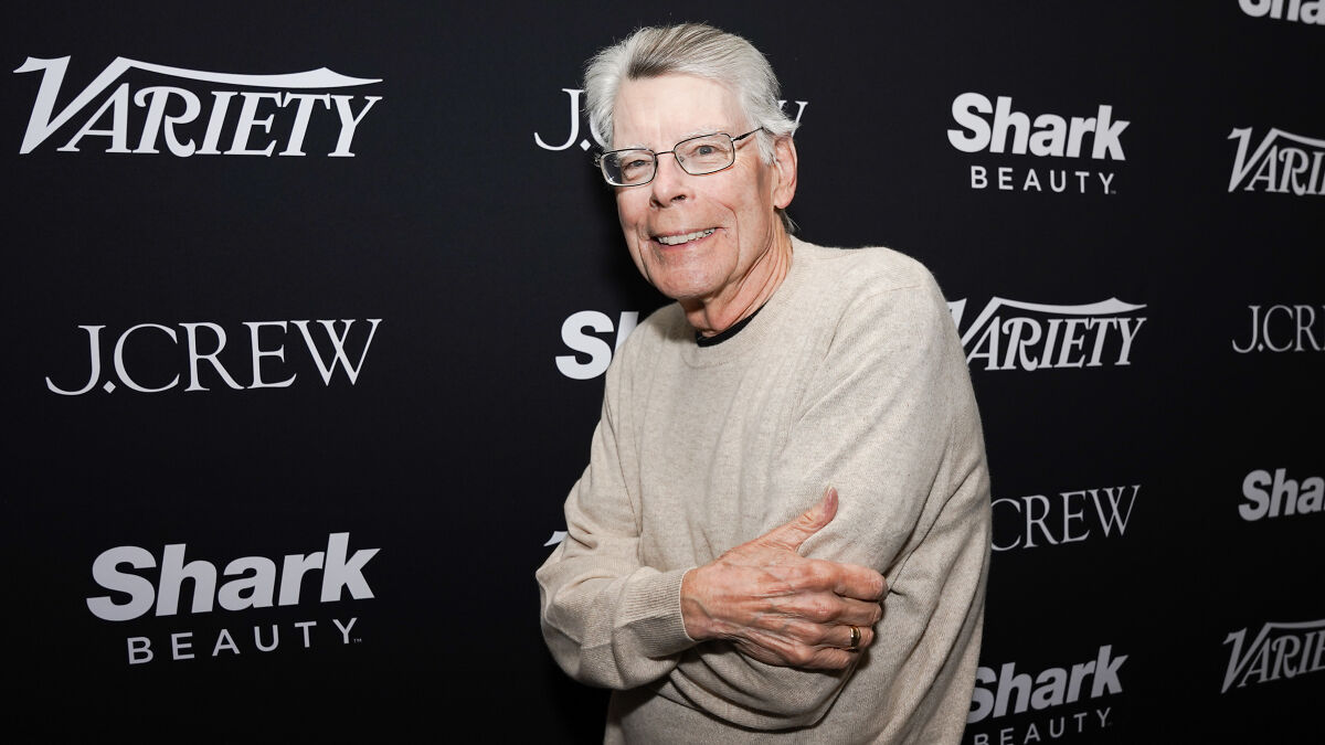 Stephen King at an event with a black backdrop featuring Variety and Shark Beauty logos, smiling and wearing glasses.