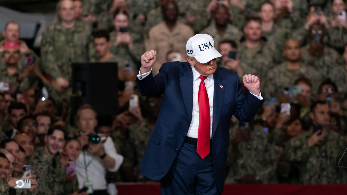 Donald Trump in a suit and USA hat, gesturing during a speech aboard an aircraft carrier with military personnel in the background.