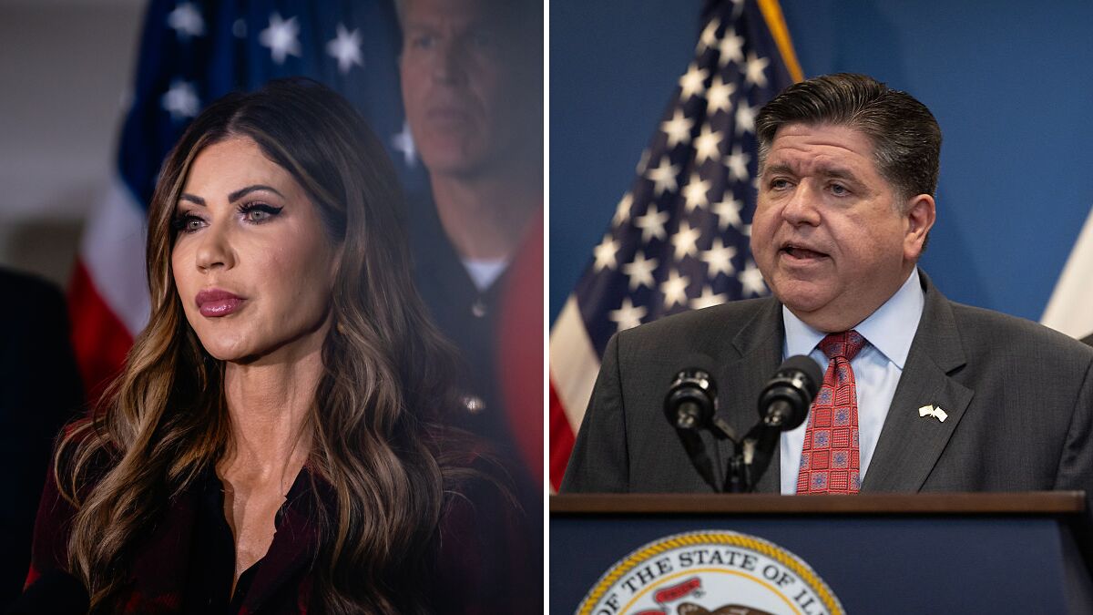 Kristi Noem speaking at a press event with the Illinois governor at a podium with American flags behind them.