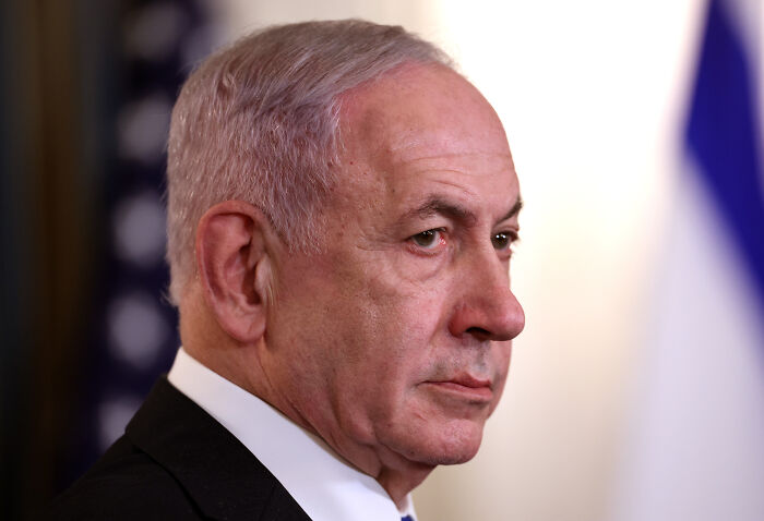 Older man in a dark suit, serious expression, in profile view with blurred American flag background, public figure context.