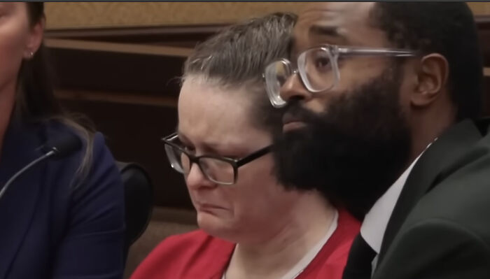 Woman with glasses in emotional distress sitting next to a man with beard and glasses during a courtroom hearing.