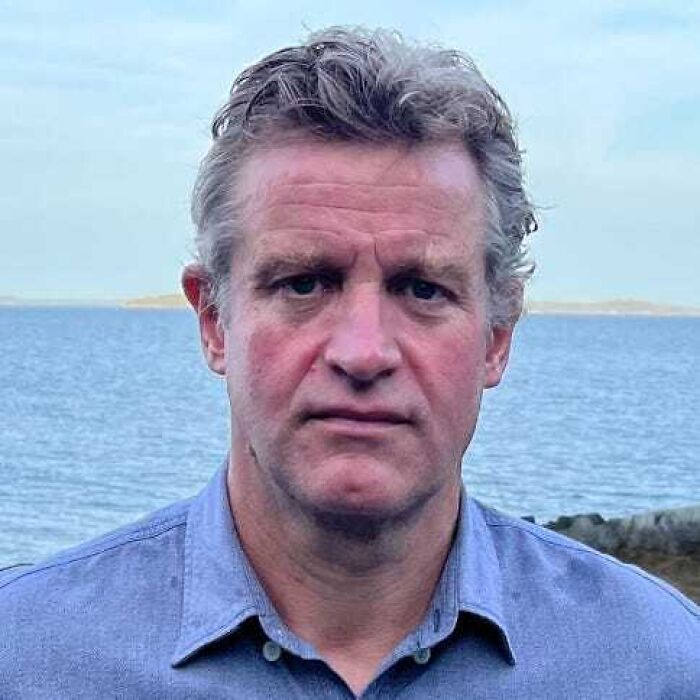 Middle-aged man with curly hair wearing a blue shirt, standing by the sea, related to influential people in Epstein’s emails.