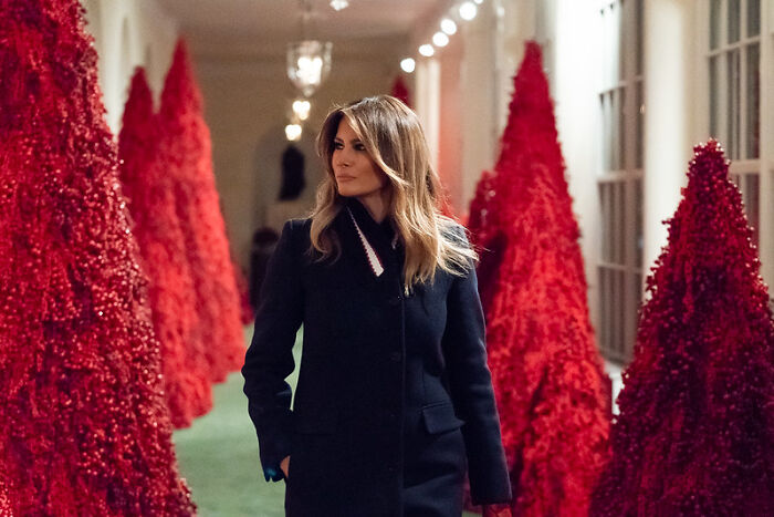 Melania Trump walking through White House hallway decorated with red Christmas trees during a holiday ceremony event.