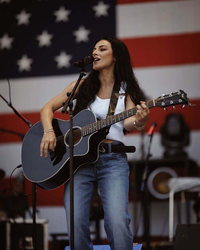 Female country musician performing on stage with guitar in front of an American flag, highlighting FBI Director Kash Patel news. Female country musician performing on stage with guitar in front of an American flag, highlighting FBI Director Kash Patel news.
