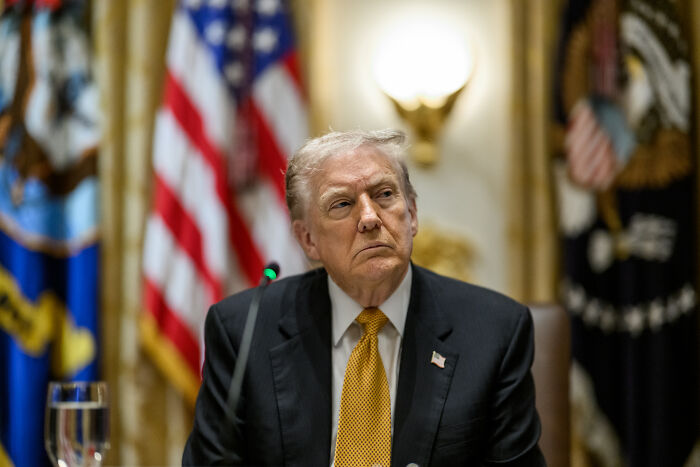 Former President Trump sitting at a meeting with American flags in the background, related to Epstein claimed Trump knew keywords.