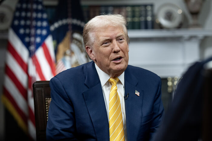 Donald Trump in a navy suit and yellow tie, speaking during an interview with American flags in the background.