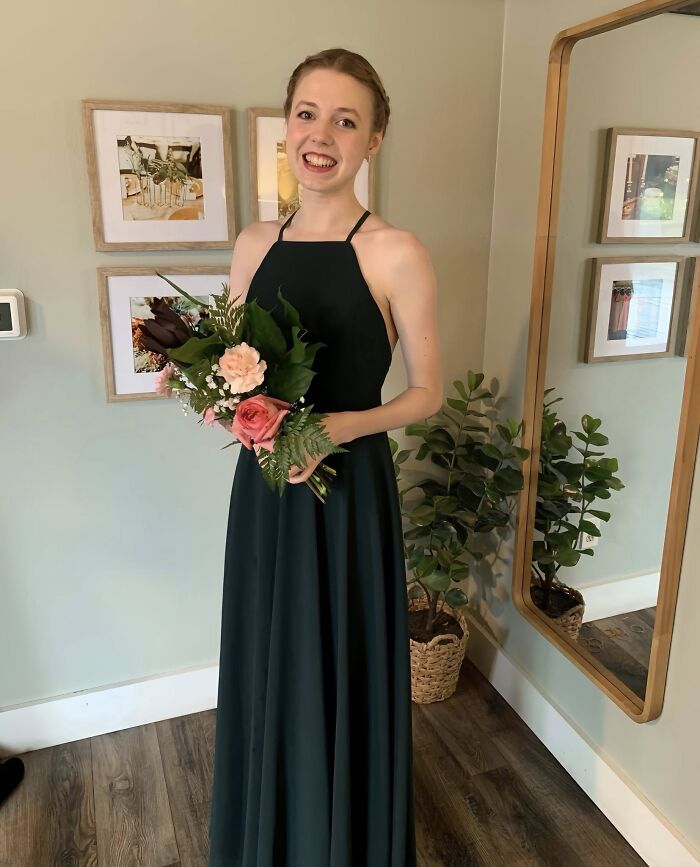 Bethany MaGee, 26, smiling in a dark green dress holding a bouquet of flowers inside a room with framed pictures.