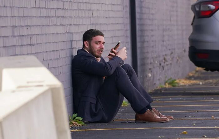 Man in tears sitting against wall outside ICE facility, holding phone after running to follow disappeared wife. Man in tears sitting against wall outside ICE facility, holding phone after running to follow disappeared wife.