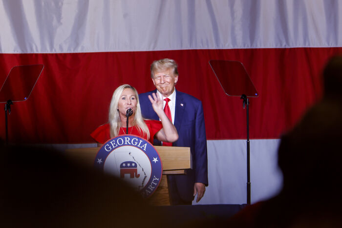 Marjorie Taylor Greene speaking at a Georgia GOP event with a large American flag backdrop and a man standing behind her.