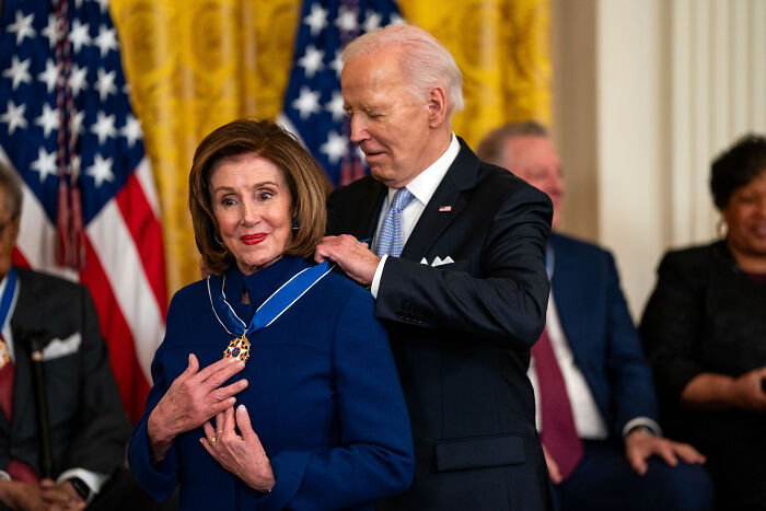 Nancy Pelosi receiving a medal from Joe Biden during a formal event with American flags in the background. Nancy Pelosi receiving a medal from Joe Biden during a formal event with American flags in the background.