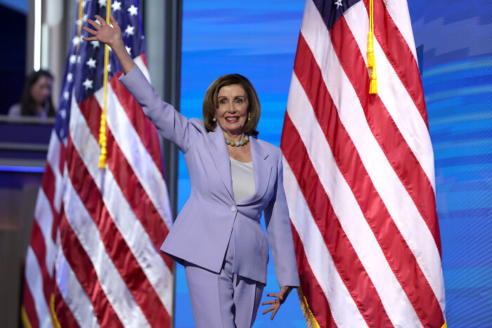 Nancy Pelosi waving on stage between American flags during announcement about not seeking re-election. Nancy Pelosi waving on stage between American flags during announcement about not seeking re-election.