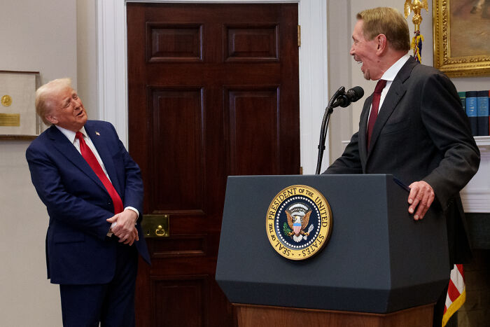 Donald Trump in a navy suit speaking with a man at a podium with the Presidential seal in a formal room Donald Trump in a navy suit speaking with a man at a podium with the Presidential seal in a formal room