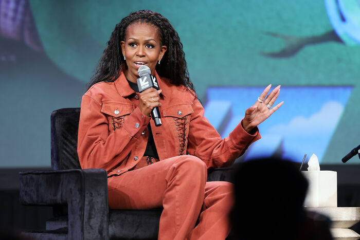 Michelle Obama speaking on stage, wearing rust-colored outfit, addressing audience about presidential run reasons. Michelle Obama speaking on stage, wearing rust-colored outfit, addressing audience about presidential run reasons.